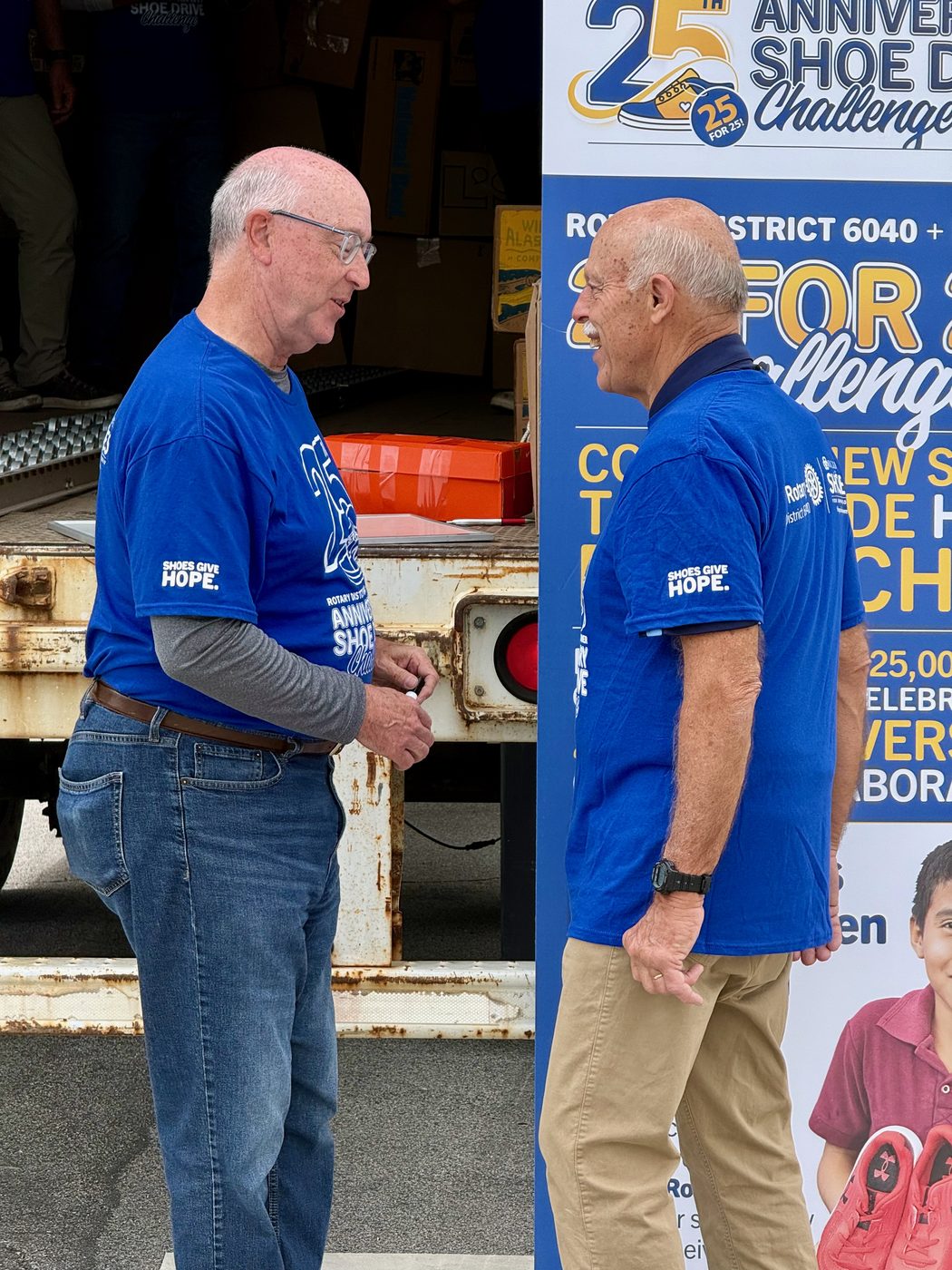 Larry Lunsford and David Rich in Rotary District 6040 shoe-drive shirts beside the 25th Anniversary banner